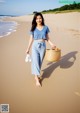 A woman walking on the beach carrying a basket and flip flops.